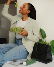 A woman with dark hair in a long braid, wearing a light green blazer and blue jeans, sits and smiles while eating green grapes from a clear container. Next to her on a white surface is a structured black top-handle handbag made of pebbled vegan leather, a clear food container lid, and a bottle of green juice.
