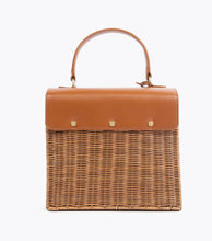 A back view of the structured picnic-style handbag. The bottom two-thirds of the bag are made of brown woven wicker, and the top flap is smooth brown vegan leather. The back of the leather flap is secured to the wicker body with three visible gold-tone fasteners.