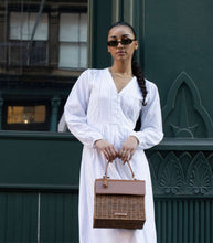 A stylish woman with braided dark hair, wearing a long-sleeved white eyelet dress and sunglasses, holds a structured handbag with a brown woven wicker body and a brown vegan leather top flap. She stands in front of a dark green door.