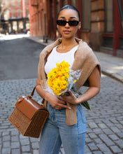 A stylish woman with hair pulled back, wearing a white tank top, a tan sweater draped over her shoulders, light-wash jeans, and sunglasses, stands on a cobblestone street. She is holding a bouquet of bright yellow flowers and carrying a structured brown wicker and leather handbag.