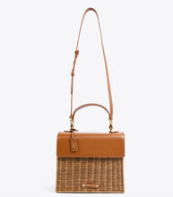 A front-facing studio shot of a two-toned handbag suspended from its thin, brown vegan leather adjustable shoulder strap. The top section is smooth brown vegan leather, and the bottom is brown woven wicker. The bag has a brown hang tag with gold "MP" initials and a gold logo plaque on the wicker.