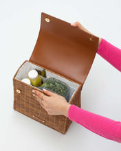 An overhead shot of the picnic-style handbag with its brown vegan leather flap held open by a hand. The body is made of brown woven wicker and features a white insulated interior. The bag is packed with two juice bottles, utensils, and a clear container holding kale.