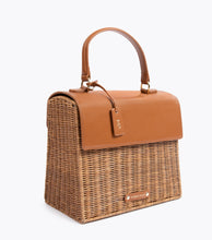 A three-quarter studio shot of a structured picnic-style top-handle bag. The lower body is made from brown woven wicker, and the top flap and handle are a contrasting smooth brown vegan leather. The bag includes a brown hang tag with gold "MP" initials and a small gold logo plaque near the bottom front.