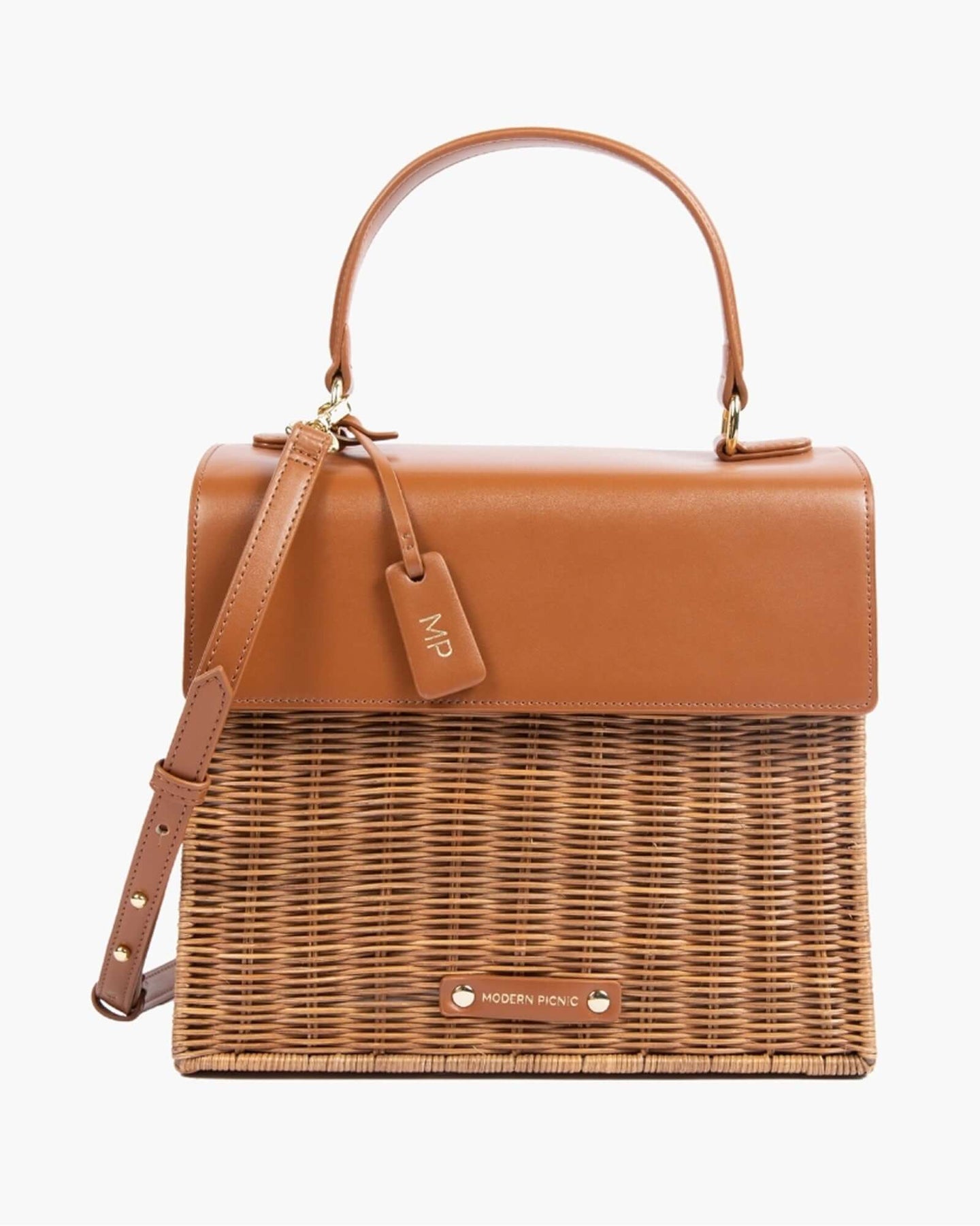 A front-facing studio shot of a structured top-handle handbag with a picnic aesthetic. The top flap, handle, and adjustable shoulder strap are smooth brown vegan leather, while the bottom half is made of brown woven wicker. The bag includes a brown hang tag with gold 