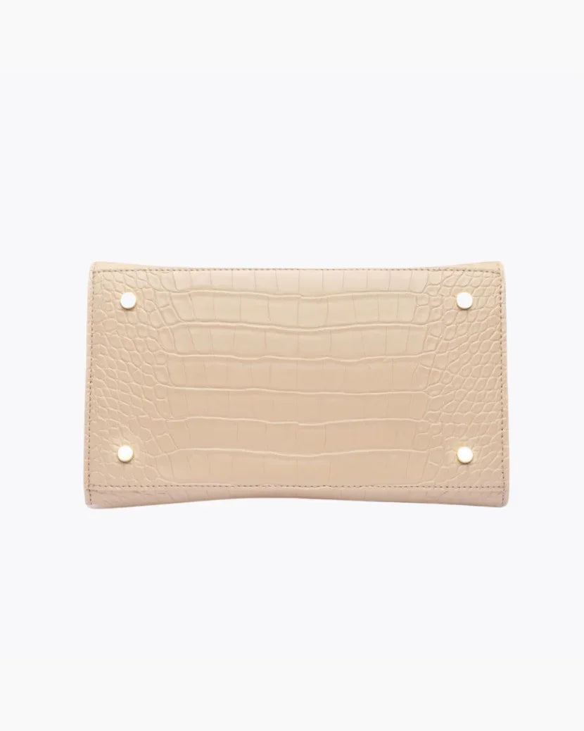 A close-up view of the bottom of the light beige handbag, showing the flat base with a crocodile-embossed texture. The base is protected by four small gold metal feet (studs).