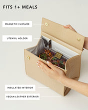 An overhead shot of a light beige handbag with a crocodile-embossed texture opened to display its function. A hand is reaching into the bag, which features a white insulated interior holding a clear container of salad. Labels point to the magnetic closure, utensil holder, insulated interior, and vegan leather exterior. The text overlay reads, "FITS 1+ MEALS."