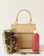A studio shot featuring a structured, light cream top-handle handbag with a crocodile-embossed texture. In front of the bag is a clear, partitioned container filled with salad greens, feta cheese, and red cabbage, alongside a bottle of pink juice and a fork.