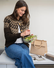 A smiling woman wearing a brown patterned cardigan and blue jeans is sitting on a white counter with a marble edge, eating a salad from a clear container. Next to her is a structured, light cream handbag with a crocodile-embossed texture and a bottle of juice.