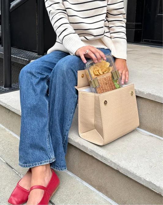 A person wearing a cream and black striped sweater, blue jeans, and red flats is sitting on stone steps. They are holding a clear lunch container with a sandwich and chips, which is resting in an open, light cream handbag that has a crocodile-embossed texture. The bag is on the step next to them, showing its gold magnetic closures.