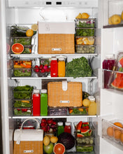 A large, well-organized refrigerator is fully stocked with clear containers of fresh produce, greens, cut fruit, and colorful juice bottles. Three versions of a small top-handle bag with a natural brown woven wicker body and a white vegan leather flap are nestled among the food containers, showcasing their compact size.