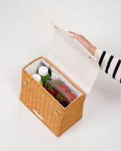 An overhead shot of the structured, smaller picnic-style handbag with its white vegan leather flap held open by a hand. The body is made of natural brown woven wicker and features a white insulated interior. The bag is packed with two juice bottles, utensils, and a clear container of fresh strawberries.