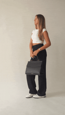 An animation shows a woman with blonde hair, dressed in a white crop top and black wide-leg trousers, modeling a structured black top-handle handbag. She carries the bag by the handle, then opens the flap to reveal the white insulated interior, showing its capacity by placing a small, clear container inside.
