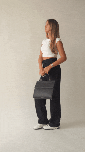 An animation shows a woman with blonde hair, dressed in a white crop top and black wide-leg trousers, modeling a structured black top-handle handbag. She carries the bag by the handle, then opens the flap to reveal the white insulated interior, showing its capacity by placing a small, clear container inside.