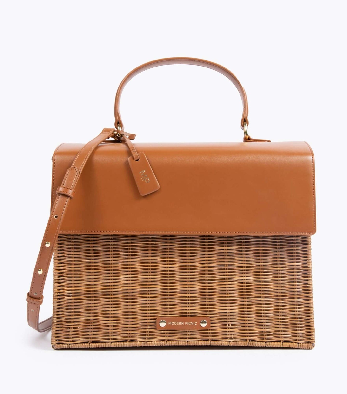 A front-facing studio shot of a structured top-handle handbag with a picnic aesthetic. The bag features a two-tone design with a smooth brown vegan leather top flap, handle, and adjustable shoulder strap, contrasted with a bottom section made of brown woven wicker. A brown hang tag with gold 