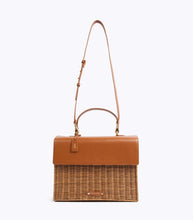 A front-facing studio shot of a two-toned handbag suspended from its thin, brown vegan leather adjustable shoulder strap. The top section is smooth brown vegan leather, and the bottom is brown woven wicker. The bag features a matching brown leather hang tag with gold "MP" initials and a small brown logo plaque on the wicker.