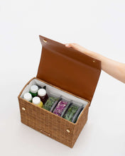 An overhead shot shows the picnic-style handbag with its brown vegan leather flap held fully open by a hand. The body of the bag is made of brown woven wicker and features a white insulated interior. The bag is packed with four juice bottles and three clear food containers holding salad ingredients like kale and purple cabbage.