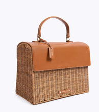 A three-quarter studio shot of a structured picnic-style top-handle bag. The lower body is made from brown woven wicker, and the top flap and handle are a contrasting smooth brown vegan leather. The bag includes a matching brown hang tag with gold "MP" initials and a small brown logo plaque near the bottom front.