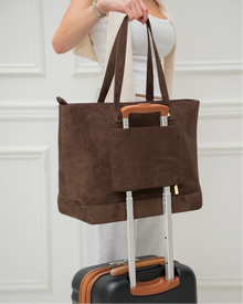 Brown suede tote bag with sleeve on luggage handle, worn by woman in white outfit