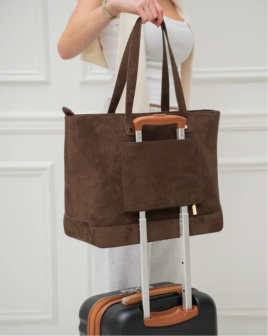 Brown suede tote bag with sleeve on luggage handle, worn by woman in white outfit