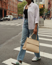 A person wearing a white button-down shirt, a black tank top, and light-wash jeans crosses a city street crosswalk. They are carrying a large, cream-colored top-handle handbag.