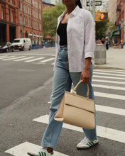 A person wearing a white button-down shirt, a black top, and light-wash jeans crosses a city street crosswalk. They are carrying a large, cream-colored top-handle handbag with a distinctive crocodile-embossed texture on the flap, and the bag features a gold top handle.