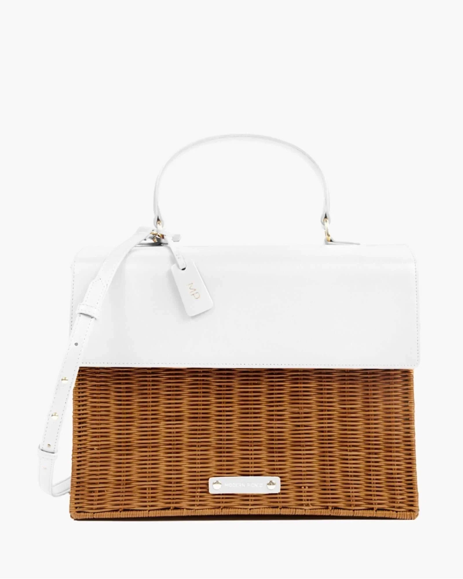 A front-facing studio shot of a unique top-handle handbag. The design is two-toned, with a smooth white vegan leather top flap and handle, and a bottom section made of brown woven wicker. A white hang tag with gold "MP" initials is attached, and a subtle logo is centered on the wicker.