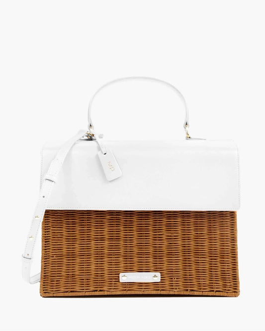 A front-facing studio shot of a unique top-handle handbag. The design is two-toned, with a smooth white vegan leather top flap and handle, and a bottom section made of brown woven wicker. A white hang tag with gold "MP" initials is attached, and a subtle logo is centered on the wicker.