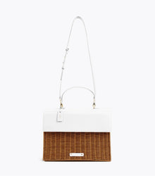 A front-facing studio shot of a two-toned handbag suspended from its thin, white adjustable shoulder strap. The top section is smooth white vegan leather, and the bottom is brown woven wicker. It features a white logo plaque, a white hang tag with gold "MP" initials, and gold-tone hardware.