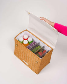 An overhead shot of the picnic-style handbag with its white vegan leather flap held open, revealing the contents. The body of the bag is made of brown woven wicker and features a white insulated interior. The bag is packed with three clear food containers holding salad greens, purple cabbage, and strawberries, along with four juice bottles.