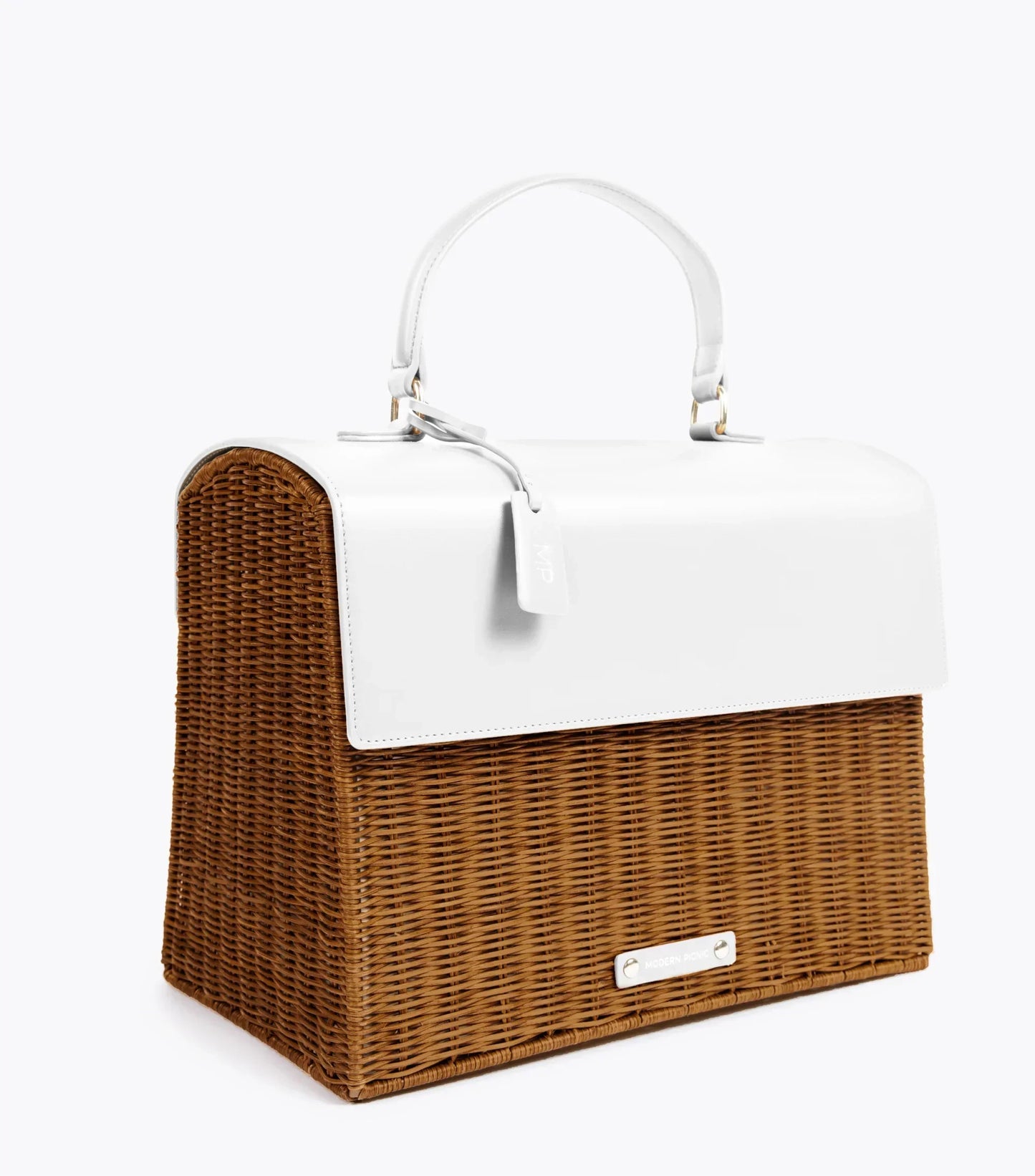 A three-quarter studio shot of a structured, vintage-inspired top-handle bag. The lower body of the bag is made from brown woven wicker, and the top flap and handle are a contrasting smooth white vegan leather. The bag includes a white hang tag and a small white logo plaque near the bottom front.