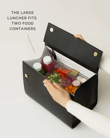 A pair of hands holds open a structured black handbag with a white insulated interior. The bag is packed with two juice bottles and one clear, rectangular food container filled with chopped vegetables. The text overlay reads, "THE LARGE LUNCHER FITS TWO FOOD CONTAINERS."
