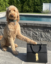 A tan Goldendoodle dog sits outside next to a pool, resting one paw on a large black handbag. The handbag features a custom portrait of the dog on its front.