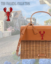 A structured handbag with a brown woven wicker body and a brown vegan leather top flap is featured against a coastal scene of quaint shingle-style houses and blooming hydrangeas. The leather flap has a hand-painted red lobster design. The text overlay at the top reads, "THE COASTAL COLLECTION."