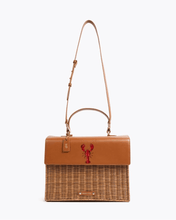 A front-facing studio shot of a two-toned handbag suspended from its thin, brown vegan leather adjustable shoulder strap. The top brown leather flap features a hand-painted red lobster design. The bottom section is made of brown woven wicker. The bag has a matching brown leather hang tag and a small gold logo plaque on the wicker.