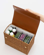 An overhead shot shows the picnic-style handbag with its brown vegan leather flap held open by a hand. The body of the bag is made of brown woven wicker and features a white insulated interior. The bag is packed with four juice bottles and three clear containers holding salad ingredients, including kale and purple cabbage.
