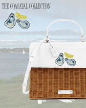 A structured handbag with a brown woven wicker body and a white vegan leather top flap is photographed outdoors against a coastal background showing a sailing ship and a bridge. The white flap features a hand-painted design of two oysters on the half shell and a lemon slice. The top text overlay reads "THE COASTAL COLLECTION."