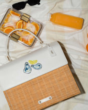 An overhead flat lay shows a picnic setup on a white blanket. The main item is a structured handbag with a natural brown woven wicker body and a white vegan leather top flap. The flap features a hand-painted design of two oysters on the half shell and a lemon slice. Picnic accessories around the bag include a pair of black sunglasses, a bottle of orange juice, and a clear container of sliced oranges.