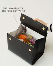 An overhead shot shows a structured black handbag with a white insulated interior held open by a hand. The bag is packed with one clear food container filled with a salad of carrots, radishes, and chopped vegetables. A fork is tucked into the side, and the text overlay reads, "THE LUNCHER FITS ONE FOOD CONTAINER."