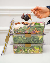 A hand holding a fork is picking up a fresh fig from the top container in a stack of three clear, rectangular food containers filled with vibrant salads. The top container features kale, carrots, crumbled cheese, and dried fruit. All containers have gold-colored locking clips.
