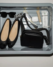 Flat lay of an airport security bin with chic travel essentials including a black Modern Picnic fanny pack, Chanel ballet flats, sunglasses, hair accessories, AirPods, lip products, and protein bars.
