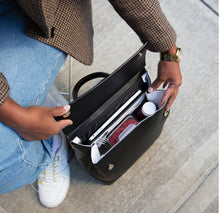 A close-up lifestyle shot showing a person in a brown blazer and jeans opening the black Modern Picnic The Backpack. The interior is packed, demonstrating the bag's function with a silver laptop in the padded sleeve, and a red food container, a white fork, and a drink bottle inside the white insulated compartment.