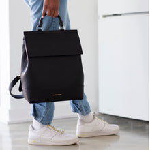 A waist-down lifestyle photo showing a person in light-wash jeans and white sneakers holding the black Modern Picnic backpack by its gold-ring top handle. The image emphasizes the bag's structured, chic silhouette and the MODERN PICNIC logo in gold at the base.