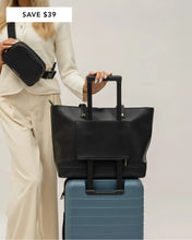 Lifestyle image of a woman traveling with a black Modern Picnic tote secured on top of a blue rolling suitcase using the trolley sleeve, while wearing a matching black Modern Picnic fanny pack across her chest. The woman is dressed in a cream sweater and matching pants, and the image features a “Save $39” promo banner in the top left corner.