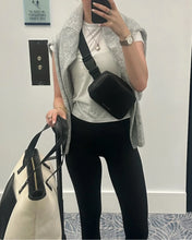 Mirror selfie of a woman holding a cream Modern Picnic tote with black accents and wearing a black Modern Picnic fanny pack crossbody, styled with black leggings, a white top, and a gray sweater for a chic travel look.