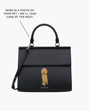 A studio shot of a structured, large black handbag made of pebbled vegan leather, featuring a hand-painted portrait of a tan Goldendoodle dog sitting on the front flap. Text on the image advertises the custom service: "SEND IN A PHOTO OF YOUR PET + WE'LL TAKE CARE OF THE REST!"