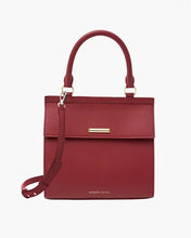 A structured red smooth luncher with a top handle with a long, removable strap, featuring a flap closure that has a small, horizontal gold-toned bar, all set against a clean white background.