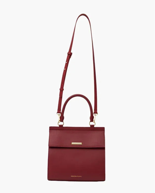 Structured red smooth luncher featuring a top handle and a long, adjustable shoulder strap is shown hanging vertically, secured with a flap closure that has a small horizontal gold-toned bar, all set against a clean white background.