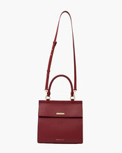 Structured red smooth luncher featuring a top handle and a long, adjustable shoulder strap is shown hanging vertically, secured with a flap closure that has a small horizontal gold-toned bar, all set against a clean white background.