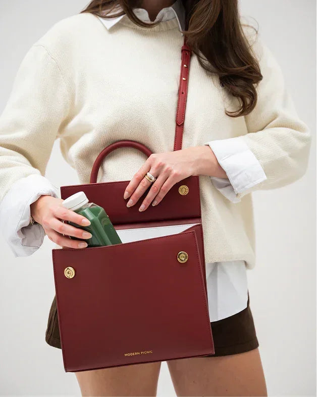 A woman in a cream sweater and white shirt holds open the red smooth lunch bag, revealing the insulated white interior as she places a green drink bottle inside, illustrating the bag's capacity for beverages.