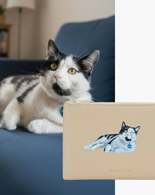 Black and white cat on sofa beside beige vegan leather pouch with cat illustration